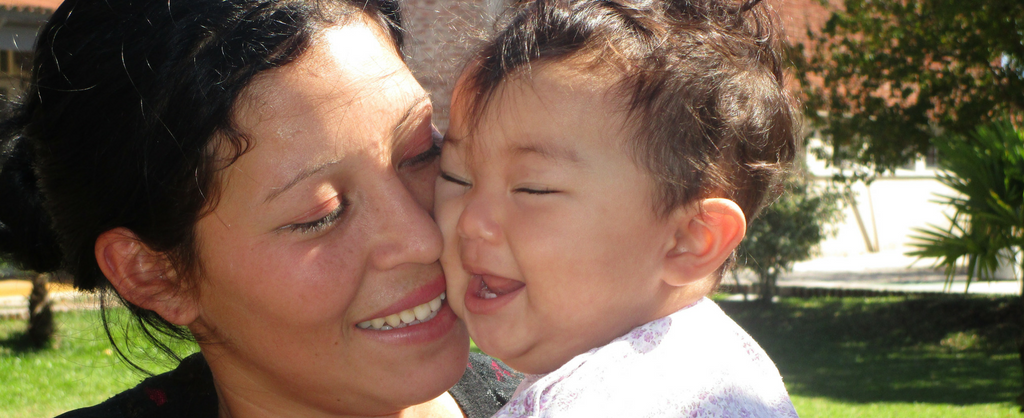 Madre sonríe mientras sostiene a su bebe cerquita de su cara. El bebé ríe.